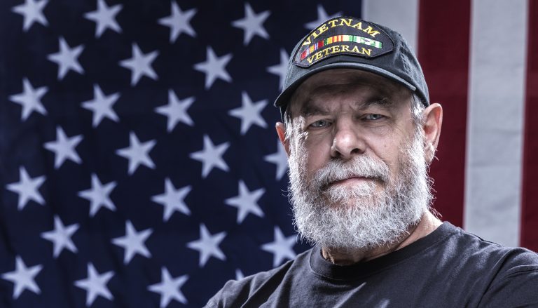 Close-up headshot of an authentic 67 year old United States Navy Vietnam War military veteran looking at the camera. Plenty of copy space on the USA flag stars and stripes red, white and blue background. He is wearing an inexpensive, non-branded, generic, souvenir shop replica Vietnam veteran commemorative baseball hat style cap.